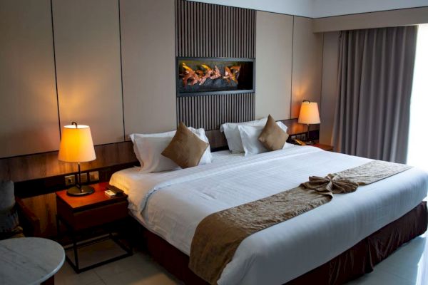 The image shows a neatly arranged hotel room with a large bed, two bedside lamps, a decorative wall feature, and a window with curtains.