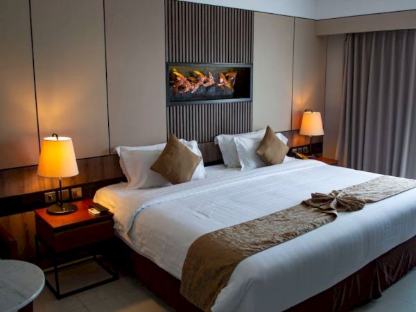 The image shows a neatly arranged hotel room with a large bed, two bedside lamps, a decorative wall feature, and a window with curtains.