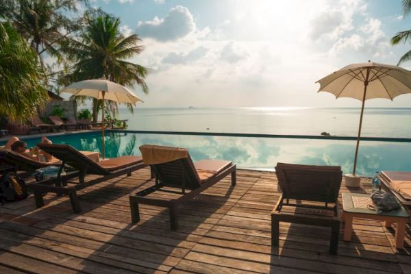 A serene poolside setting with lounge chairs, umbrellas, and a stunning view of the ocean under a partly cloudy sky and setting sun.