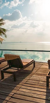 A serene poolside scene with lounge chairs, umbrellas, palm trees, and an infinity pool reflecting the sky and ocean beyond, under morning sun.