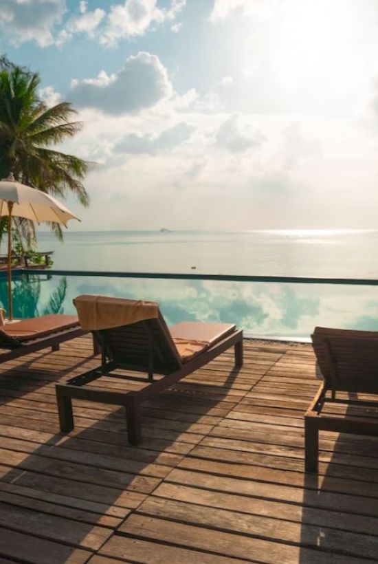 A serene poolside scene with wooden lounge chairs, umbrellas, and palm trees overlooking calm waters under a partly cloudy sky.