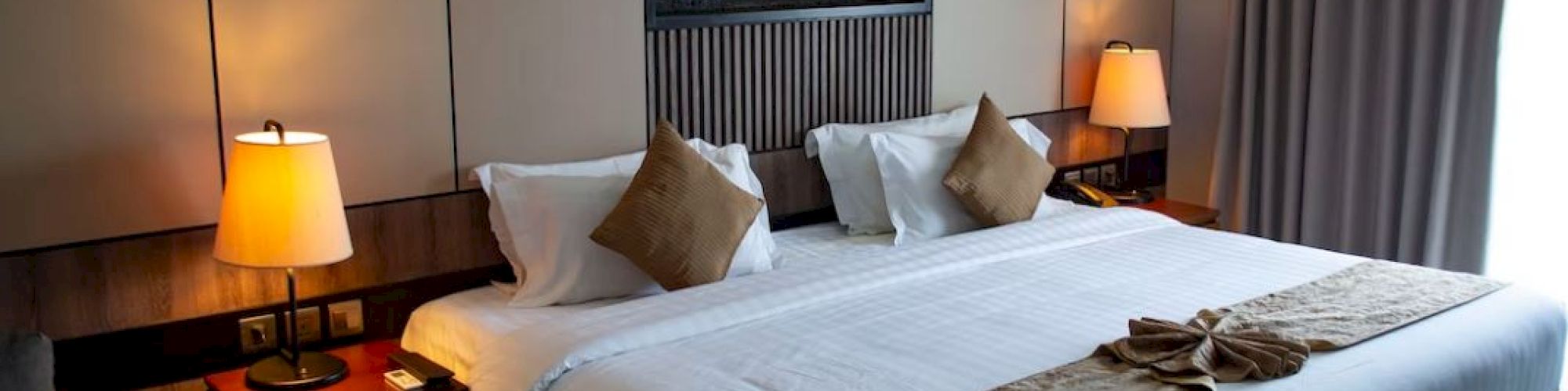 A well-lit hotel room features a king-sized bed with brown pillows, bedside tables with lamps, and a decorative artwork above the bed.