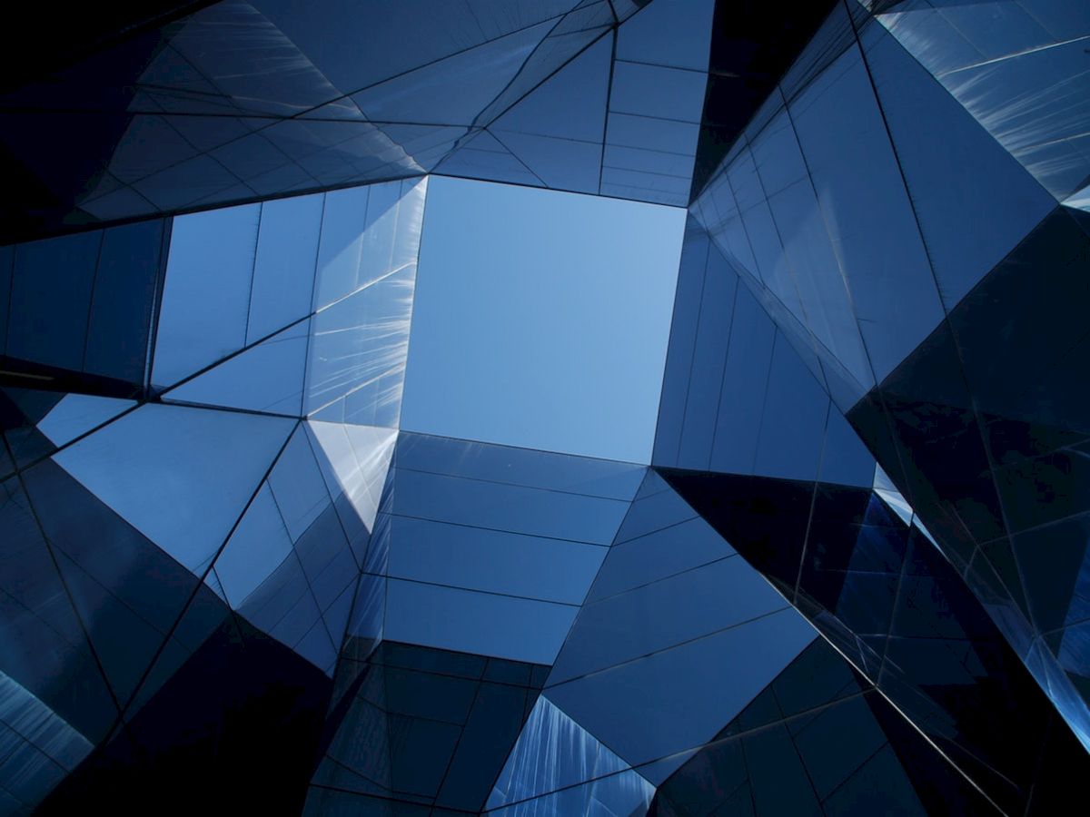 The image shows an upward view of a modern building with reflective glass panels and geometric shapes, creating a visually striking pattern.
