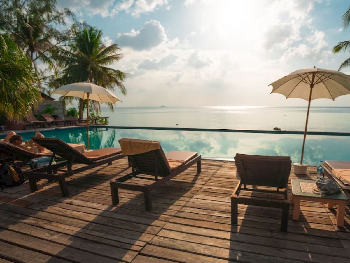 The image shows a tropical poolside setting with lounge chairs, umbrellas, and palm trees, overlooking a calm body of water under a blue sky.