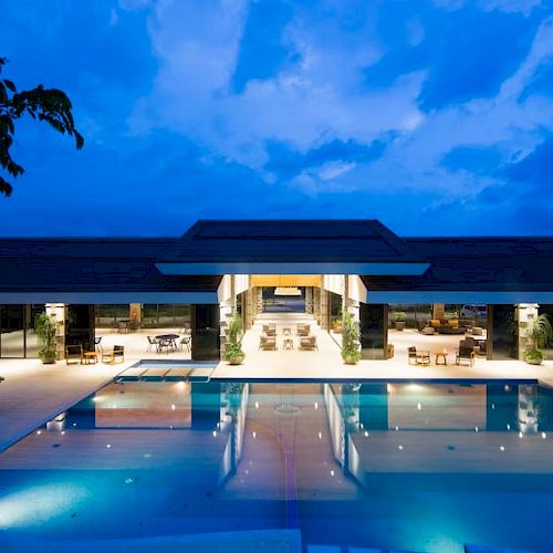 A luxurious outdoor pool area in front of a modern, well-lit building at dusk, with clear blue skies and trees in the background.