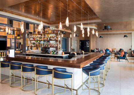 A modern bar with blue barstools, pendant lights, and a fully stocked back bar. Lounge seating is visible in the background.