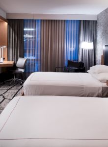The image shows a modern hotel room with two beds, a flat-screen TV, a desk, and a large window with blinds, featuring a clean and contemporary design.
