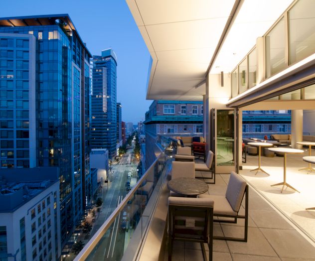The image shows a modern balcony with seating, overlooking a cityscape with tall buildings at dusk.