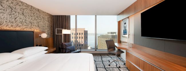 A modern hotel room features a large bed, wall-mounted TV, desk with chair, and floor-to-ceiling windows offering a city view with buildings.