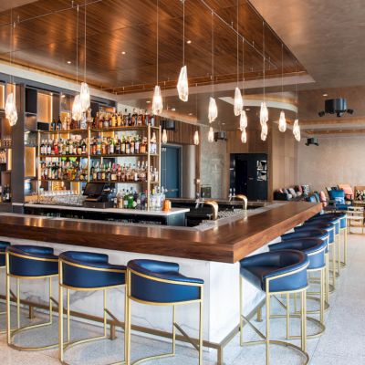 A modern bar with a wooden counter, blue bar stools, hanging lights, and a fully stocked shelf of bottles, situated in a spacious, stylish room.