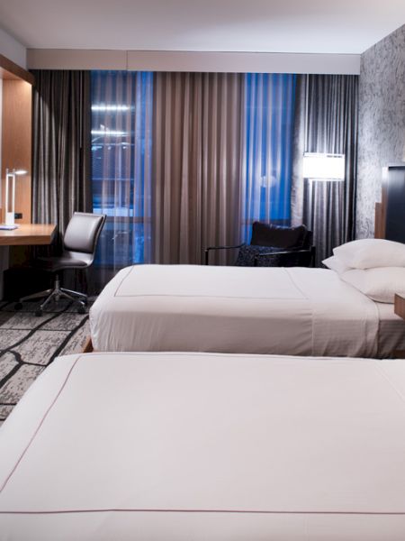 The image shows a modern hotel room with two neatly made beds, a flat-screen TV, a work desk, a chair, and a window with curtains.
