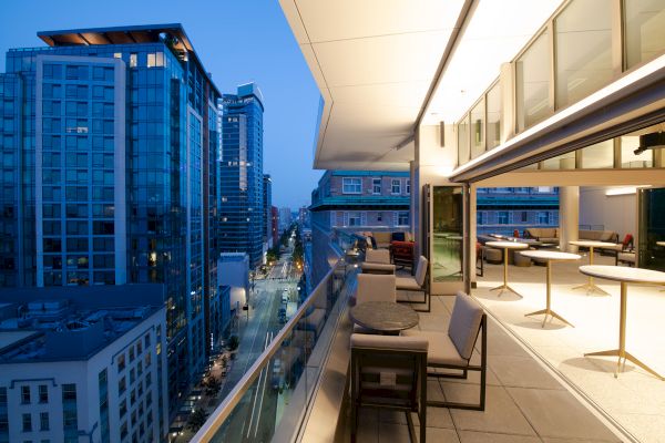 The image shows a balcony with tables and chairs overlooking a city at dusk, with tall buildings and a street visible below.