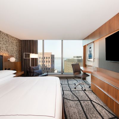 A modern hotel room with a large bed, a work desk with a chair, a flat-screen TV, and a window offering a city and water view.