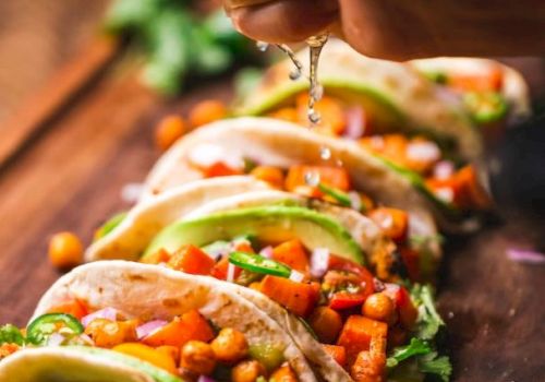 The image shows a person squeezing a lime over a row of tacos filled with chickpeas, diced vegetables, avocado slices, and garnished with herbs.