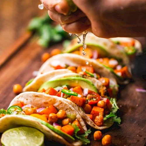 The image shows a person squeezing a lime over a row of tacos filled with chickpeas, diced vegetables, avocado slices, and garnished with herbs.