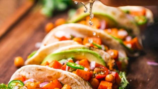 The image shows a person squeezing a lime over a row of tacos filled with chickpeas, diced vegetables, avocado slices, and garnished with herbs.