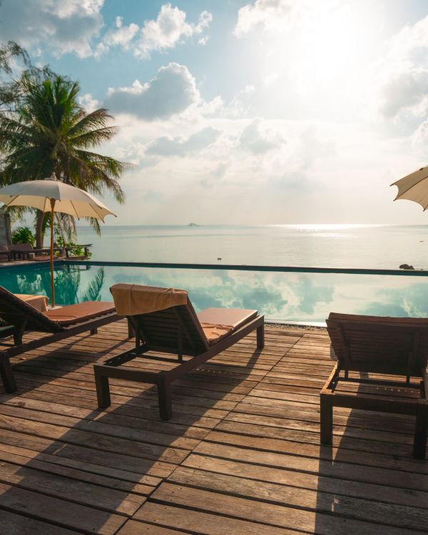 The image shows lounge chairs and umbrellas by a pool overlooking the ocean with a backdrop of palm trees and a partially cloudy sky.
