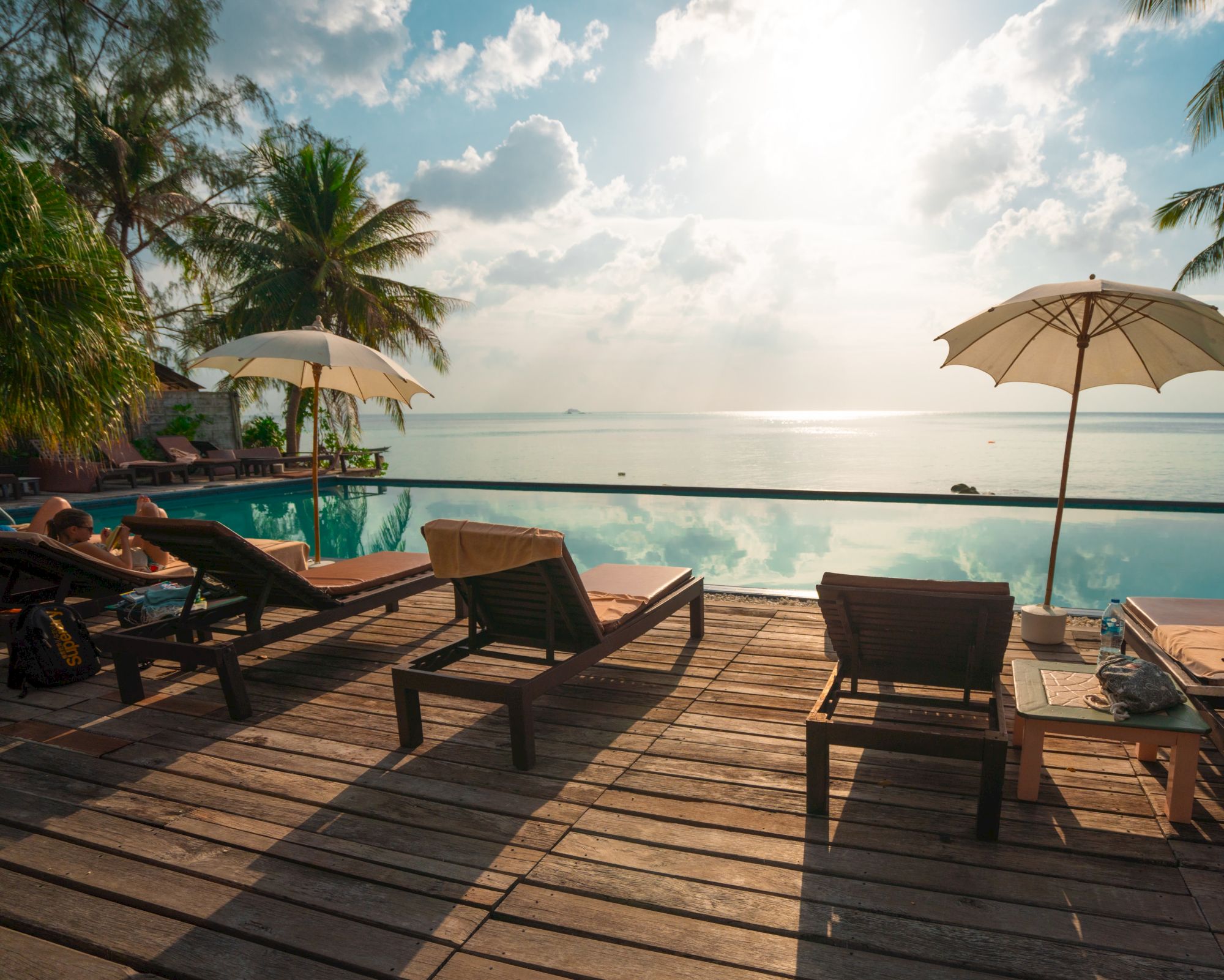 A serene tropical setting with lounge chairs, umbrellas, and a pool overlooking a calm ocean during sunset, surrounded by lush palm trees and greenery.