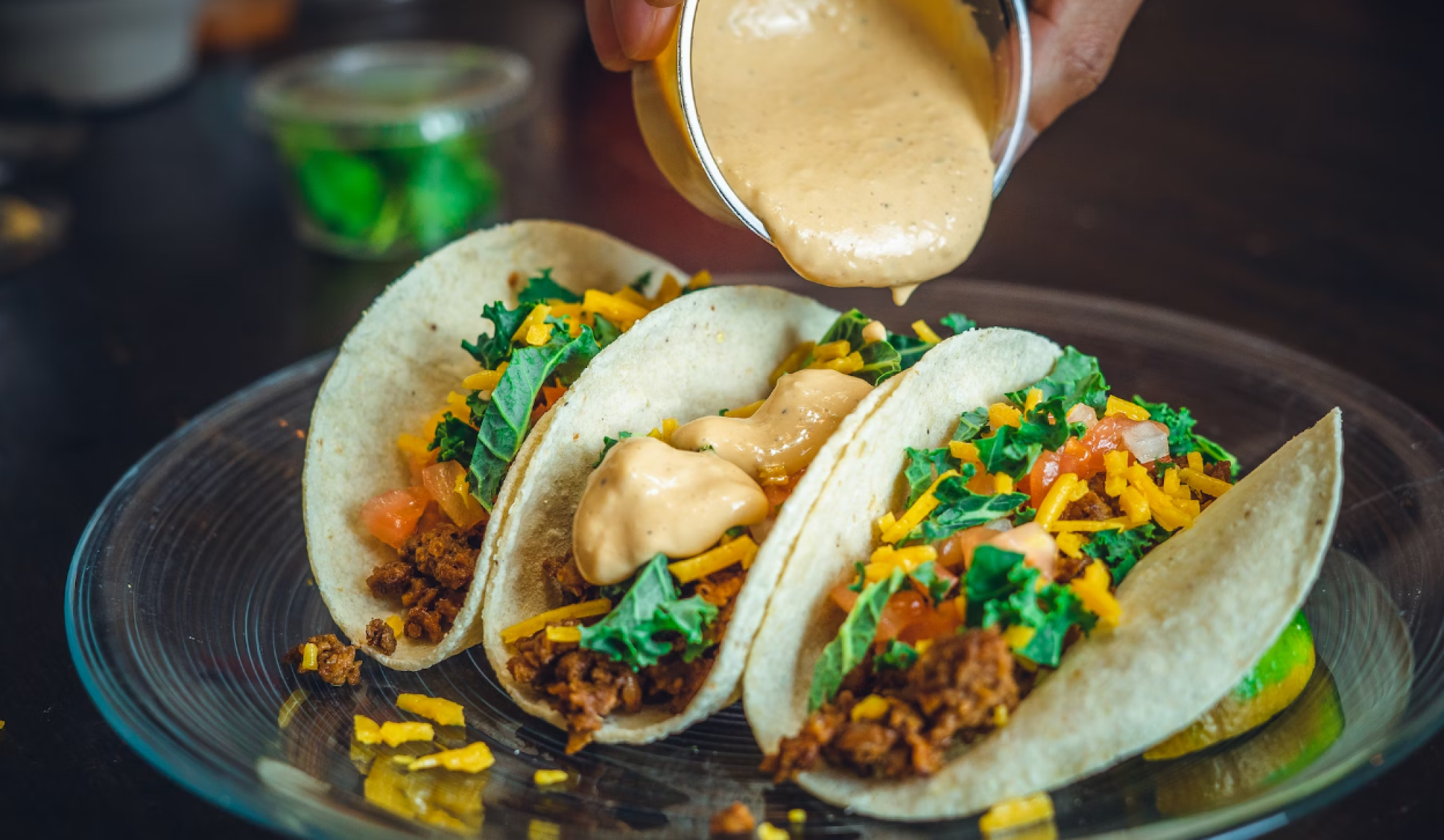 A person pouring sauce onto three tacos filled with ground meat, shredded cheese, tomatoes, and greens on a glass plate.