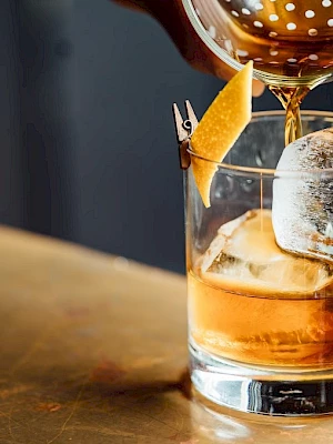 A drink is being poured into a glass with ice and a lemon peel garnish on a wooden surface.