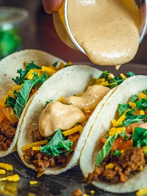 The image shows three tacos filled with meat, greens, cheese, and tomatoes on a plate, with cheese sauce being poured on them.