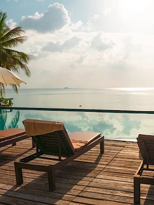 Wooden deck with loungers, umbrellas, an infinity pool, and a scenic view of the ocean under a partly cloudy sky. Palm trees are in the background.
