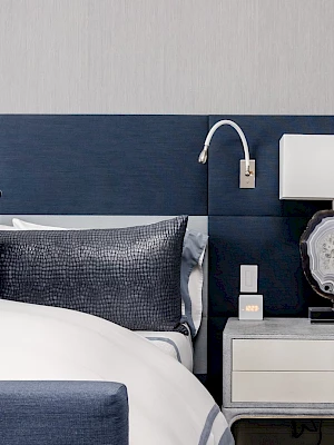 The image depicts a modern bedroom with a navy blue headboard, matching pillows, a light grey nightstand, and a stylish lamp on a wooden side table.