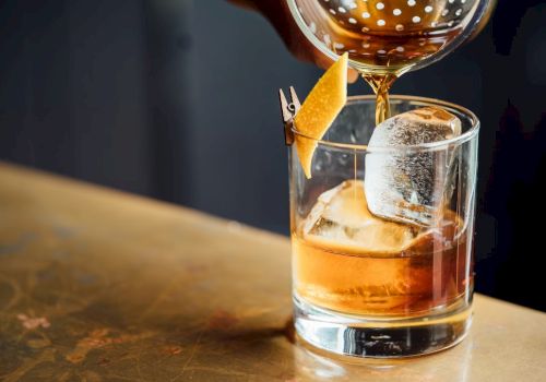 A hand is pouring a cocktail from a strainer into a glass with ice and a citrus peel garnish on a bar counter.