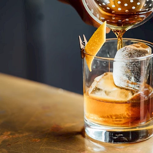 A hand is pouring a cocktail from a strainer into a glass with ice and a citrus peel garnish on a bar counter.