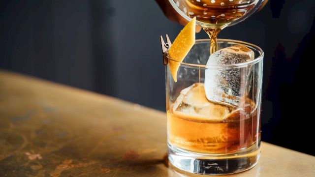 A hand is pouring a cocktail from a strainer into a glass with ice and a citrus peel garnish on a bar counter.