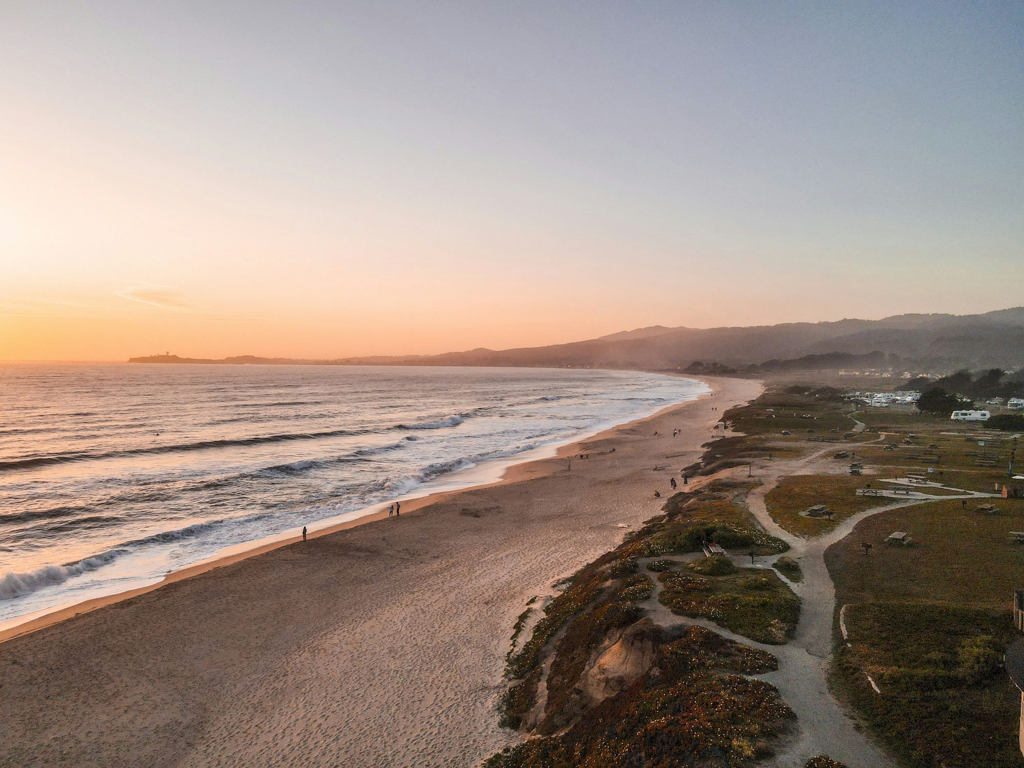 This image shows a serene beach at sunset with gentle waves, a sparsely populated shoreline, and a coastal landscape with paths and grassy areas.