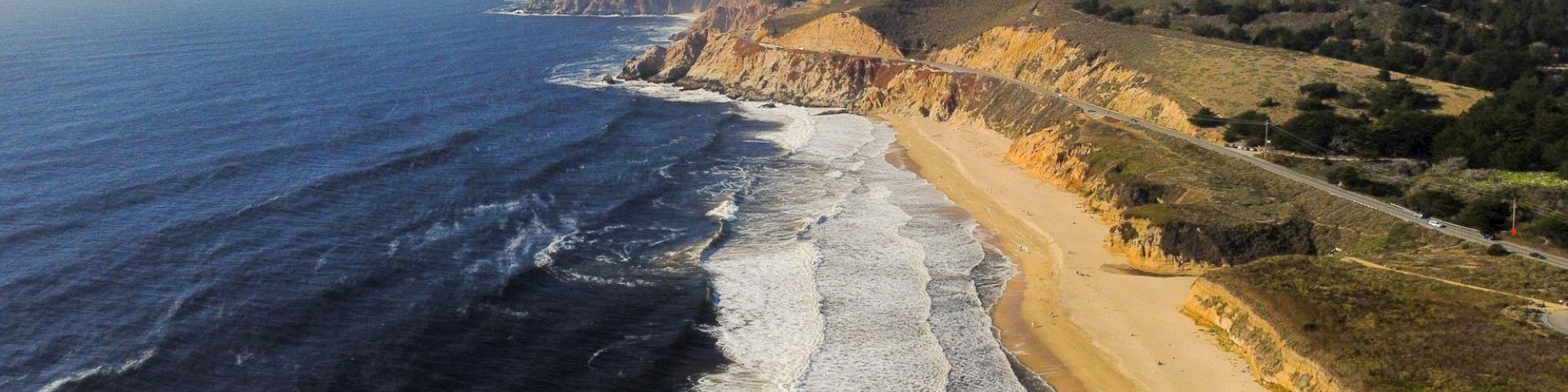 An aerial view of a coastal landscape with waves crashing onto a sandy beach, rugged cliffs, and rolling hills in the background.