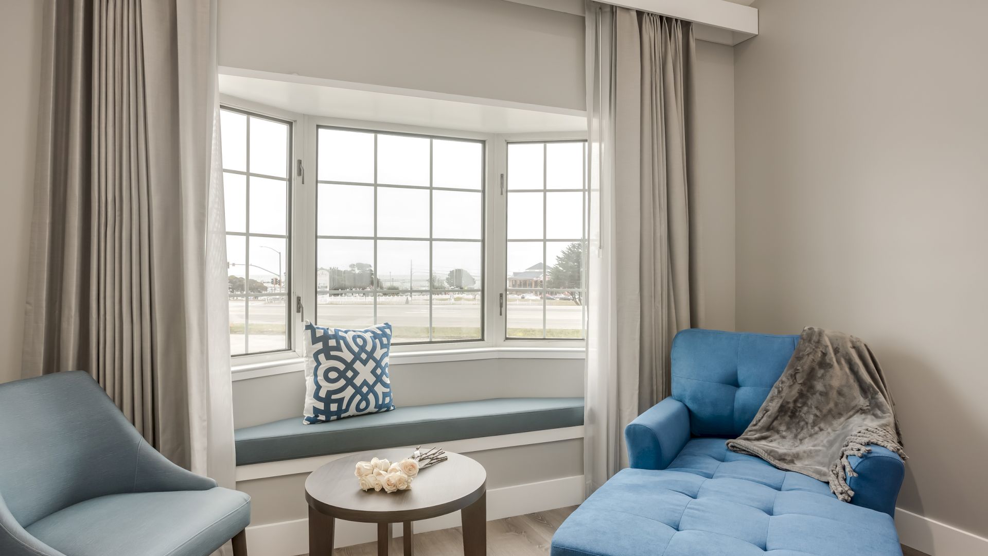 A cozy sitting area with a blue chaise lounge, a blue chair, a small round table, and a window seat with a cushion by a large window.