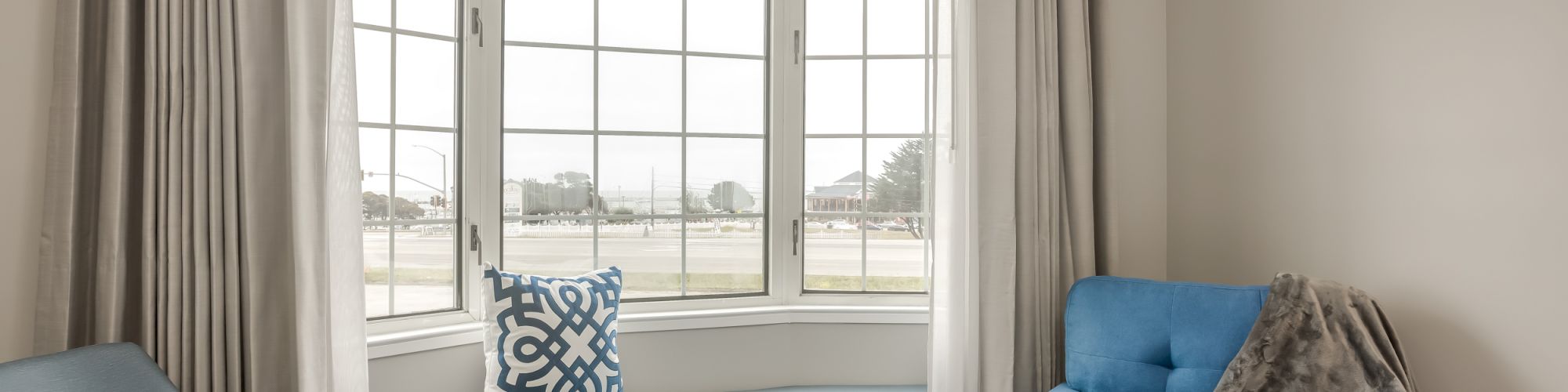 A cozy sitting area with a blue chaise lounge, a blue chair, a small round table, and a window seat with a cushion by a large window.