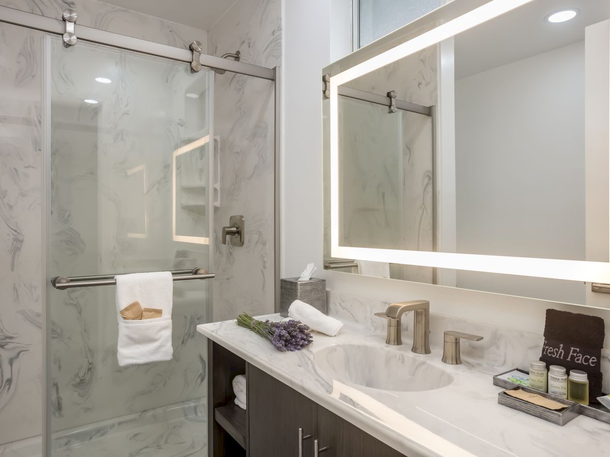 A modern bathroom with a marble shower, glass door, illuminated mirror above a sink, towel and toiletries neatly arranged on the counter, and lavender sprigs.