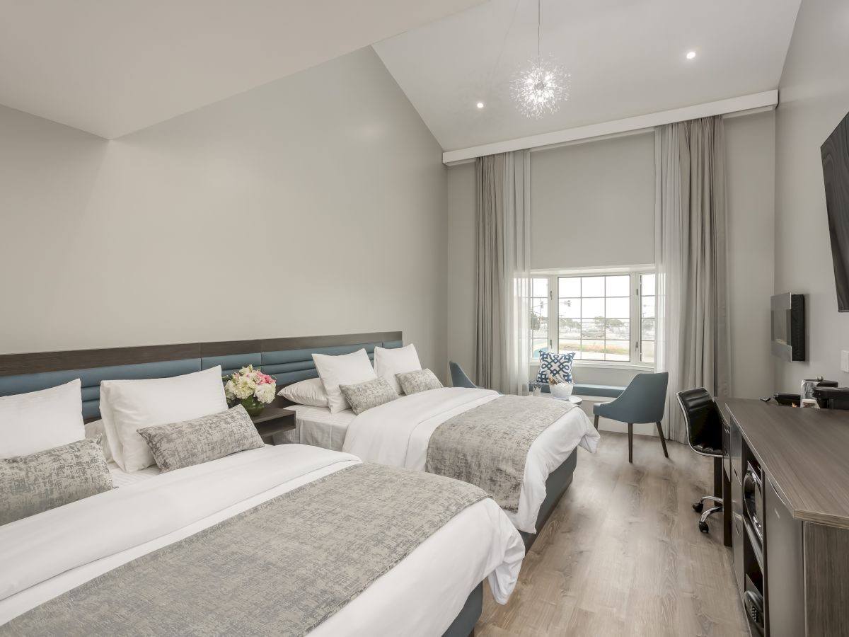 The image shows a modern hotel room with two double beds, a window with curtains, a desk, a chair, a TV, and light decor on the wall.