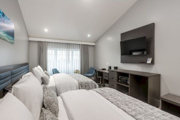 A modern hotel room with two neatly made beds, a TV mounted on the wall, a desk, microwave, and a window with curtains at the far end of the room.