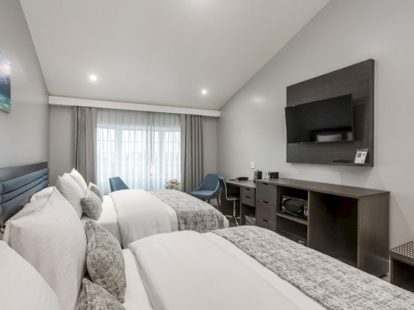 A modern hotel room with two neatly made beds, a TV mounted on the wall, a desk, microwave, and a window with curtains at the far end of the room.