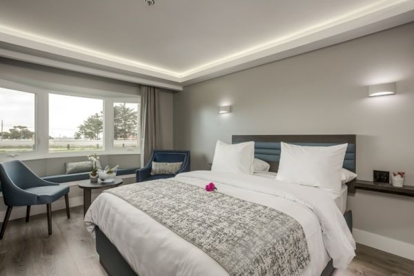 A modern hotel room with a neatly made bed, two blue chairs, a small table, and large windows looking out to a scenic view, and soft lighting.