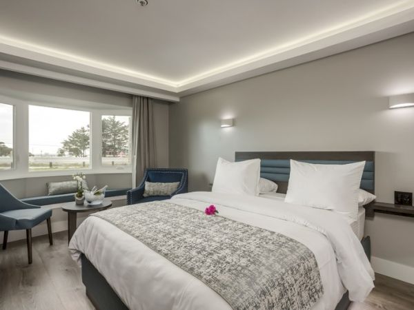 A modern hotel room with a neatly made bed, two blue chairs, a small table, and large windows looking out to a scenic view, and soft lighting.