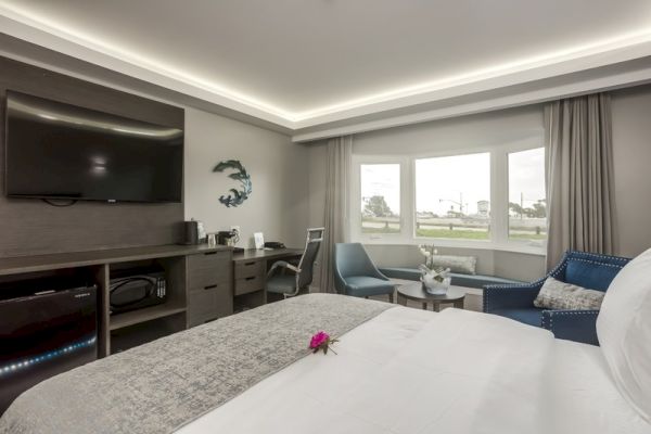 A modern hotel room features a large bed, television, desk, chair, and window with a view. Elegant decor, including a flower, adds a touch of charm.