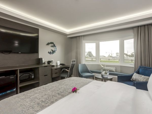 A modern hotel room features a large bed, television, desk, chair, and window with a view. Elegant decor, including a flower, adds a touch of charm.