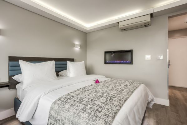 A modern hotel room features a neatly made bed with white linens, a wall-mounted air conditioner, and a flat-screen TV.