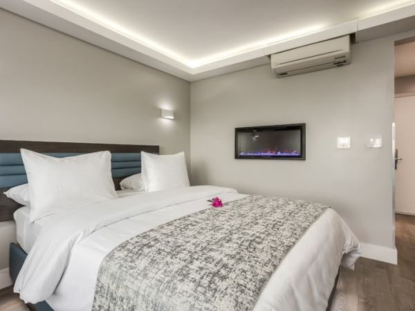 A modern hotel room features a neatly made bed with white linens, a wall-mounted air conditioner, and a flat-screen TV.
