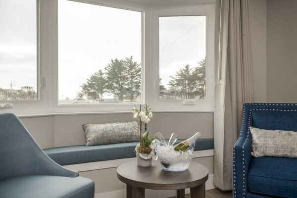 A cozy seating area with blue chairs, a table with ice bucket and champagne, small potted plant, large windows, and a serene outdoor view.