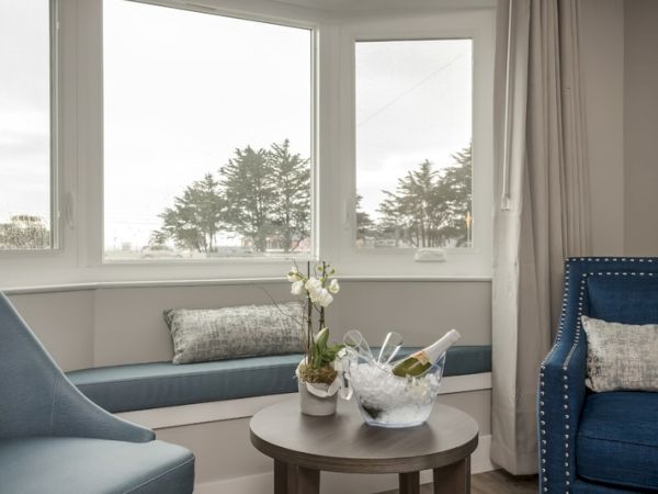 A cozy seating area with blue chairs, a table with ice bucket and champagne, small potted plant, large windows, and a serene outdoor view.
