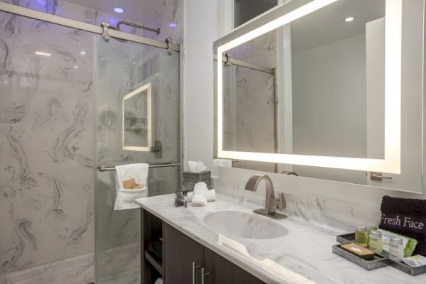 A modern bathroom with a marble countertop, lighted vanity mirror, walk-in glass shower, fresh towels, and assorted toiletries on the counter.