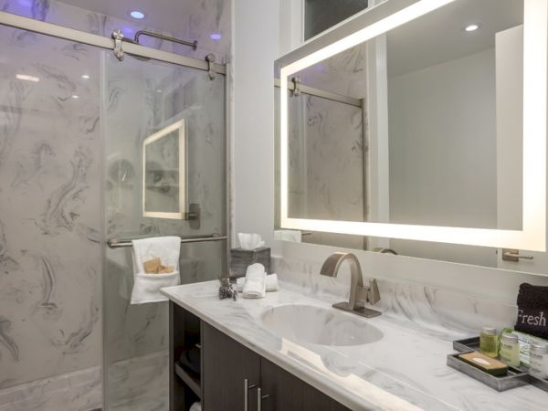 A modern bathroom with a marble countertop, lighted vanity mirror, walk-in glass shower, fresh towels, and assorted toiletries on the counter.