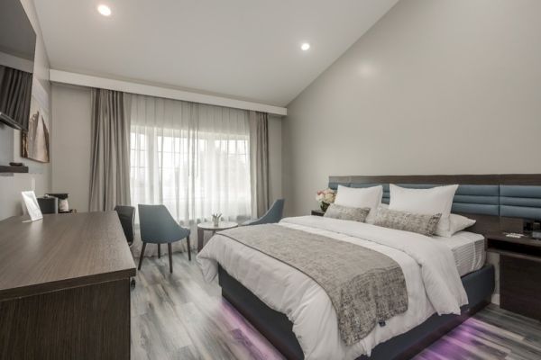 A modern bedroom featuring a large bed, two chairs with a small round table, a desk, and large windows with curtains, in a minimalist style.