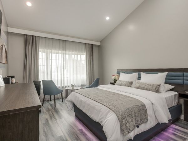 A modern bedroom featuring a large bed, two chairs with a small round table, a desk, and large windows with curtains, in a minimalist style.